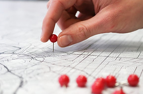 A hand places a red pushpin on a detailed map, with several other red pushpins lying nearby on the map surface.