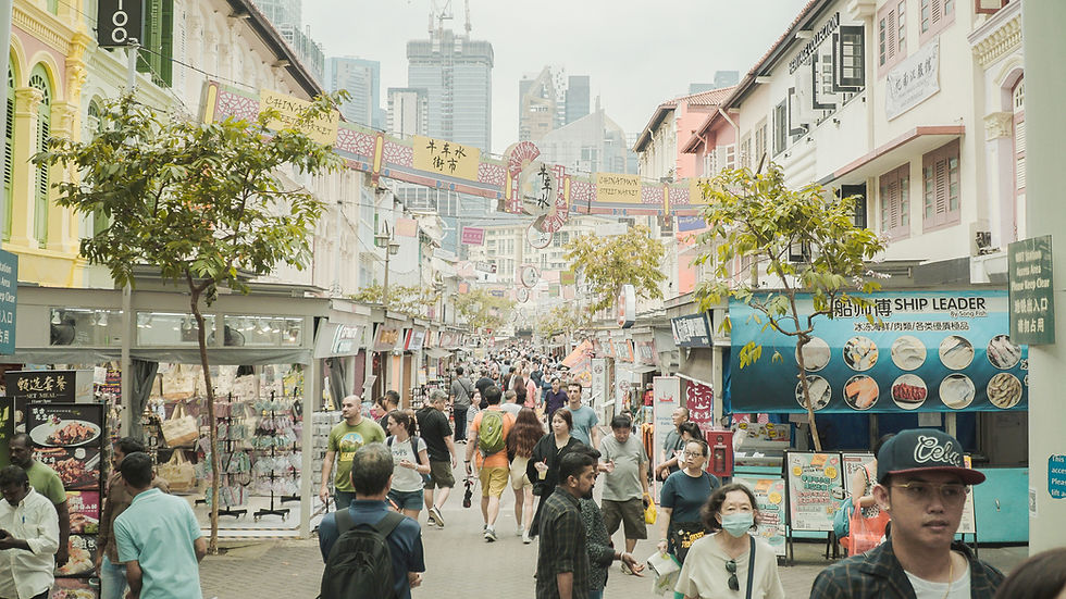 Crowded Chinatown street market with shoppers, colorful shop signs, and city skyscrapers rising in the hazy background.