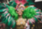 A woman wears elaborate dark green and pink feather wings and head-dress, plus a purple pink and yellow flower crown