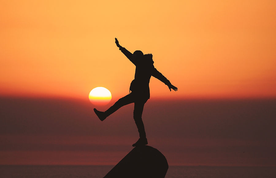 A person balancing on one leg on a rock at sunset. It looks like the sun is a ball balanced on there foot like a football representing the fine balance of our divine masculine and feminine.