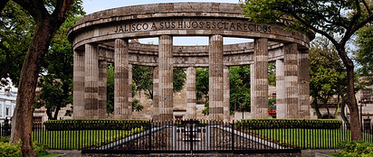 Circular stone monument with columns, surrounded by trees and greenery. The inscription reads "Jalisco a sus hijos esclarecidos." A fenced area with a small garden is in front of the monument.