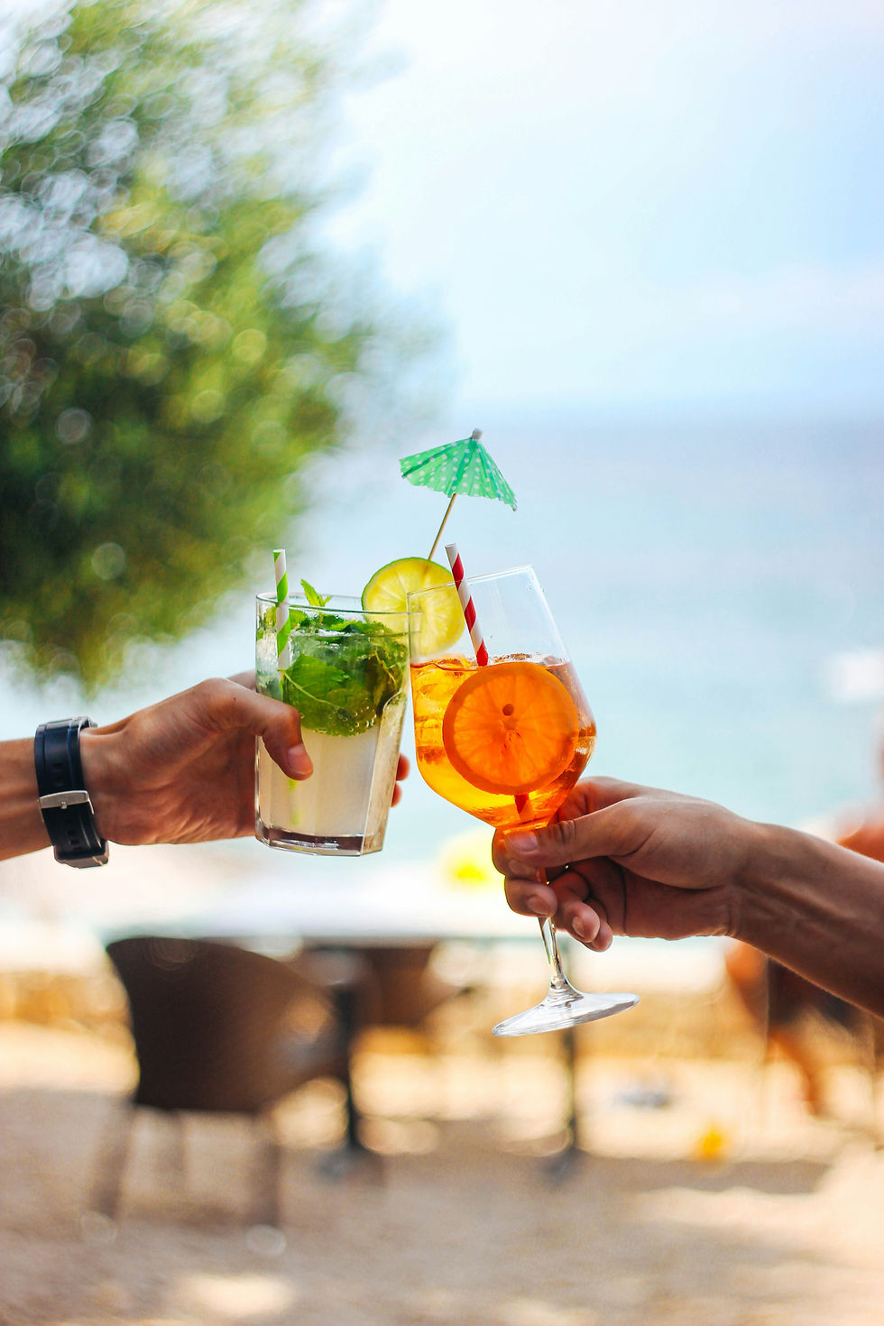 Two hands toast a mojito and orange spritz with fruit garnish and mini umbrellas at a sunny outdoor café.