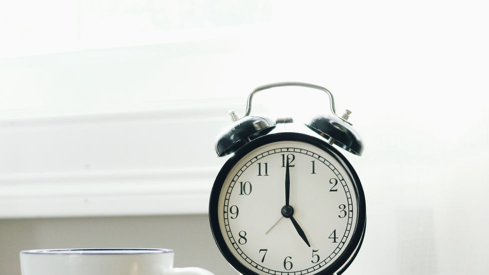 Black alarm clock on stacked books with "Still Waters." White mug with black stripes nearby, on a wooden surface. Bright background.