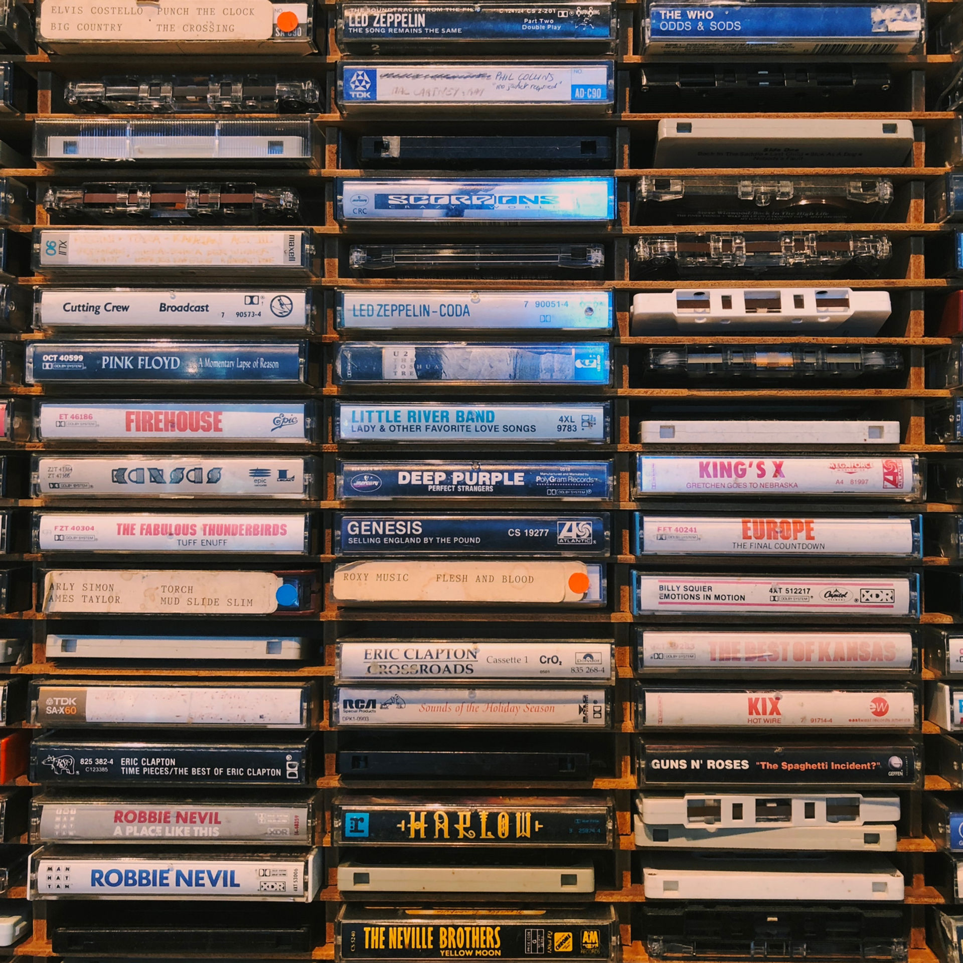 Wall of stacked cassette tapes in wooden shelves, labeled with rock bands like Led Zeppelin, Pink Floyd, and Genesis.