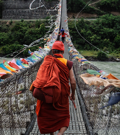 Buddhist monk in traditional robes walking across a suspension bridge in Bhutan, surrounded by lush green valleys and misty mountains