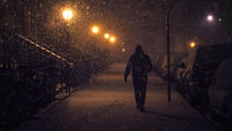 A man walking down a sidewalk at night. The lamplights are glowing.