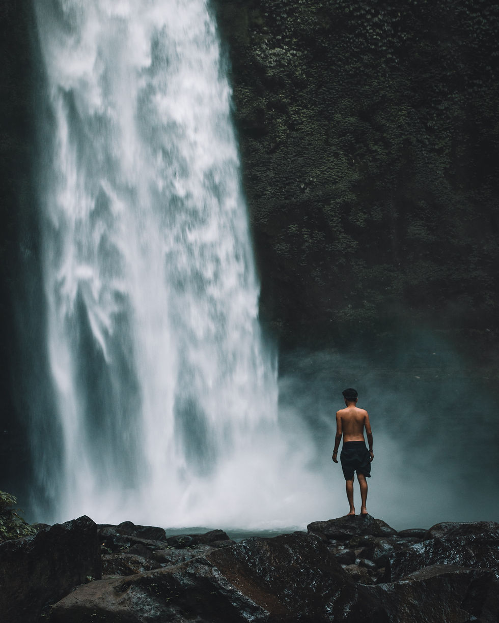A lone traveler standing in front of a powerful Bali waterfall, surrounded by mist and lush greenery.