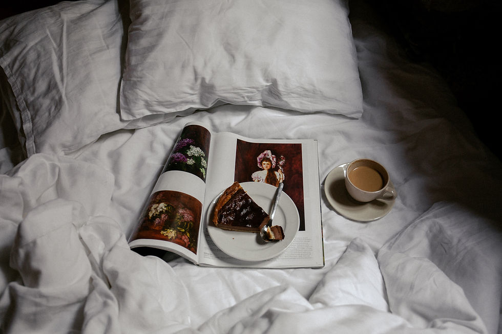 Slice of pie on an open magazine with floral images, next to a cup of coffee on a white bed. Cozy and relaxed atmosphere.