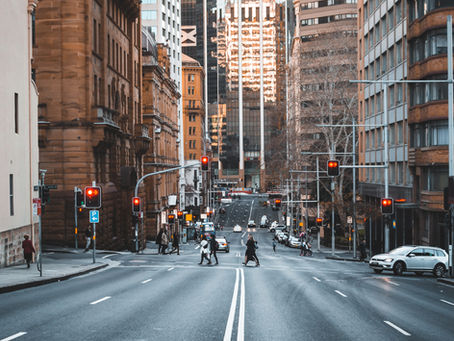 City street with pedestrians crossing at a red-light intersection. Tall buildings and parked cars line the road. Traffic volume and pedestrian movement in urban setting