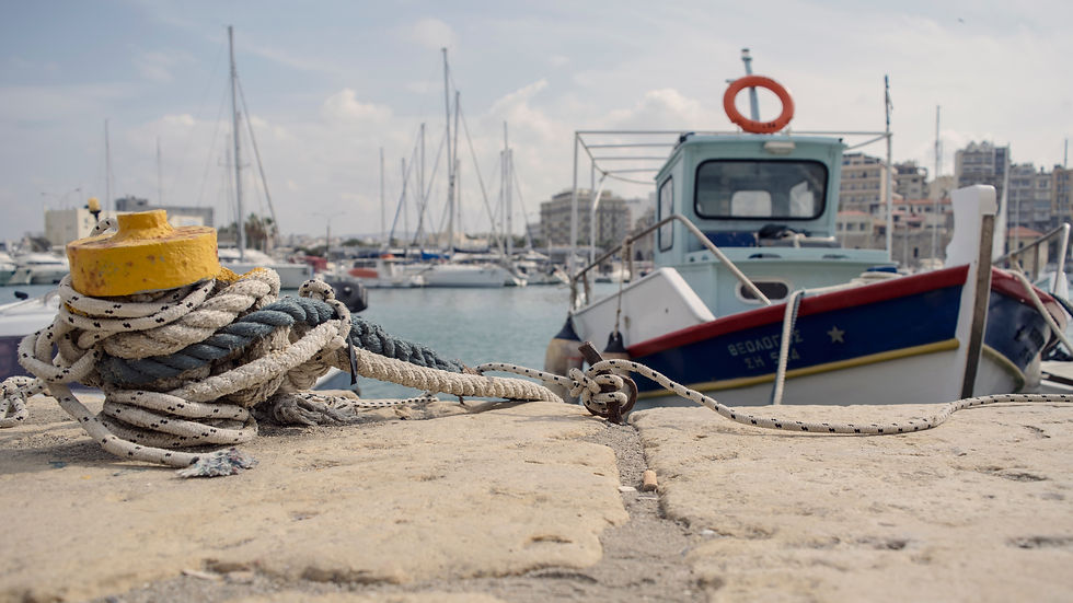 A traditional fishing boat moored with thick ropes at the old Venetian harbor in Heraklion, Crete, with sailboats and the city skyline blurred in the background.