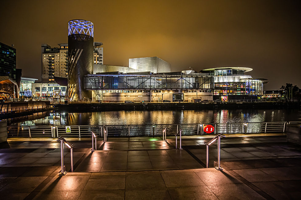 Modern waterfront building illuminated at night, reflecting on the water. Stairway and railing in foreground, under a cloudy sky.