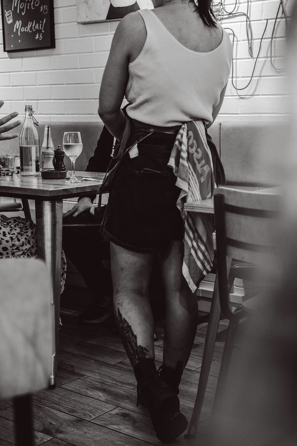 Waitress in apron stands by a restaurant table with water and wine, chalkboard menu reading Mojito $13 and Mocktail $8.