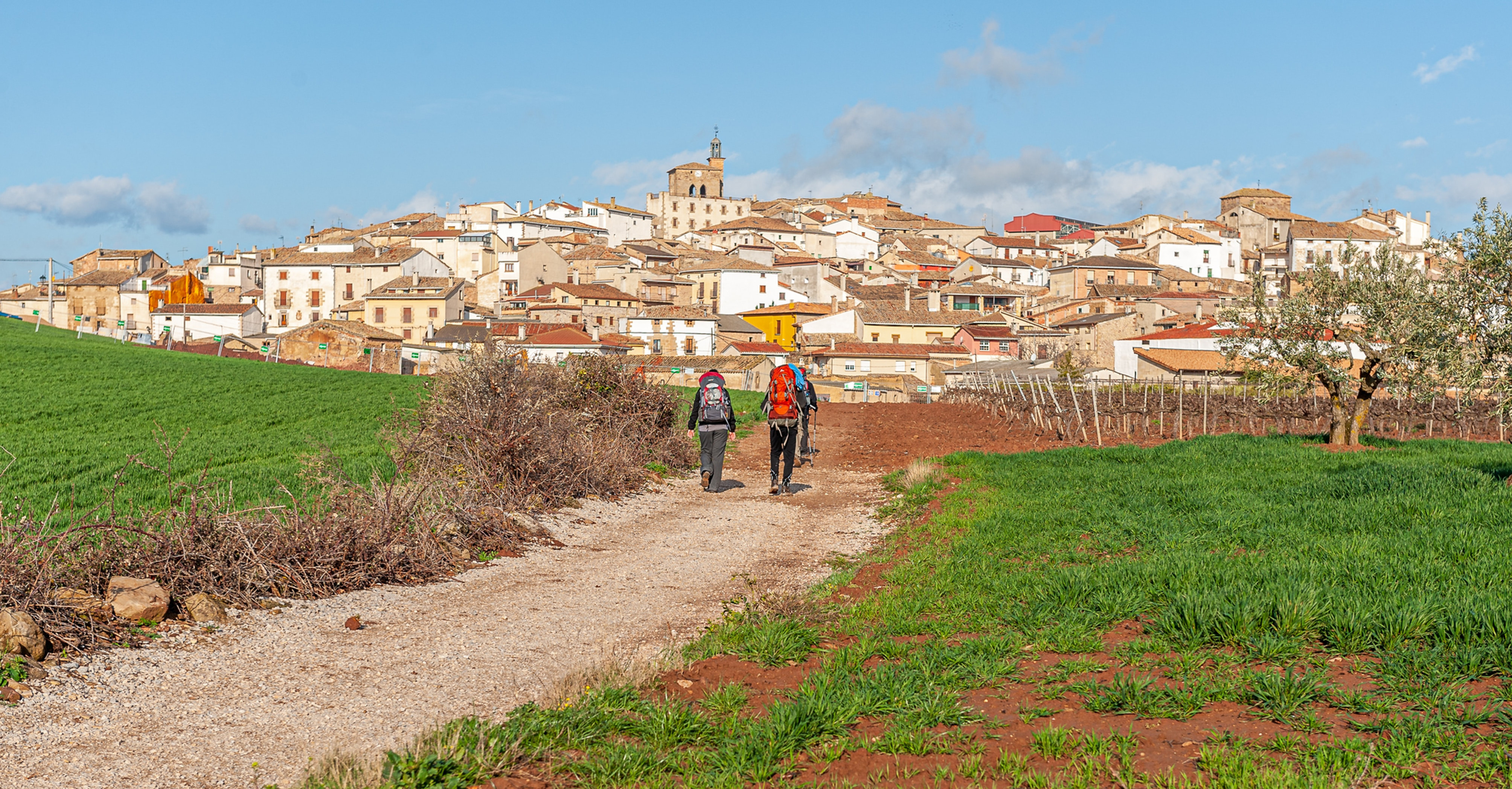 Two hikers with backpacks walk a gravel path toward a hilltop village, with green fields and a blue sky overhead.