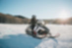 Person riding a snowmobile on a snowy field with trees in the background. Sun shining brightly, creating a clear, crisp atmosphere.