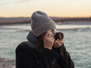 A person in a grey knit hat photographs a riverside sunset, wearing a dark coat. The sky is pastel, creating a serene ambiance.