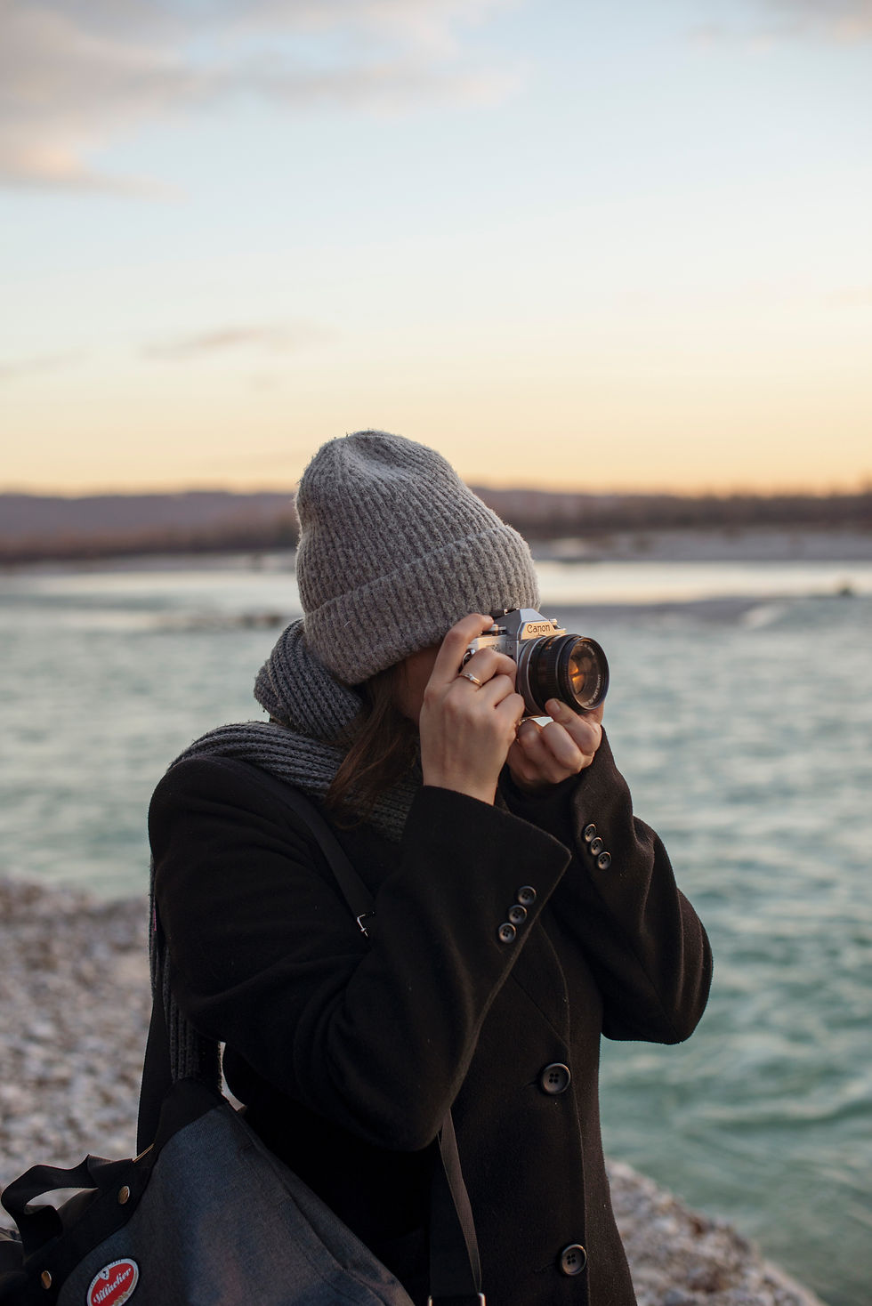 A person in a grey knit hat photographs a riverside sunset, wearing a dark coat. The sky is pastel, creating a serene ambiance.