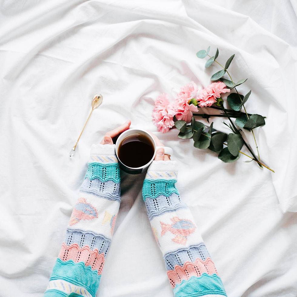 A woman's arms clothed in knitted pastel jumper clutch a coffee mug on a white tablecloth next to a spray of pink flowers.