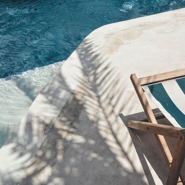 Striped deck chair beside a blue pool; palm shadows cross the sunlit stone. Relaxing, serene summer vibe.