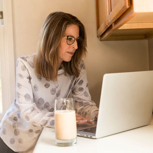 Woman in glasses working on a laptop in a kitchen, wearing a white patterned blouse. A glass of smoothie is on the counter. Bright, calm setting.