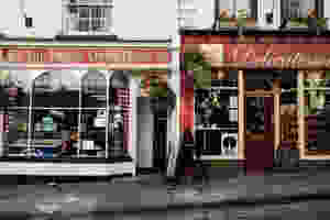 A British Quintessential Retail High Street sccne of Two shop fronts, one with a Red Sign above the window reading: The Priory Coffee House. Next door to the right, is a Store with a Sign Saying about the Door Delecatessen. The Shops are on a slight inclide to the right and the road surface is cobble. It is summer as hanging baskets are hanging from the store fronts.
