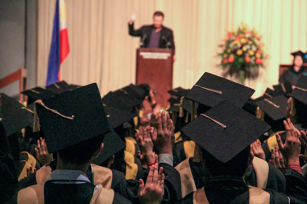 Graduates in caps face a speaker at a podium. A flag and flowers are visible in the background, creating a celebratory atmosphere.