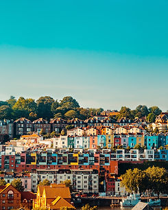 A view from across a river of 5 rows of houses, tiered down a hill. Each house is painted in a different block colour: green, orange, yellow, turqoise, red and white. There is a row of trees at the top of the hill, and the sky above is clear blue.
