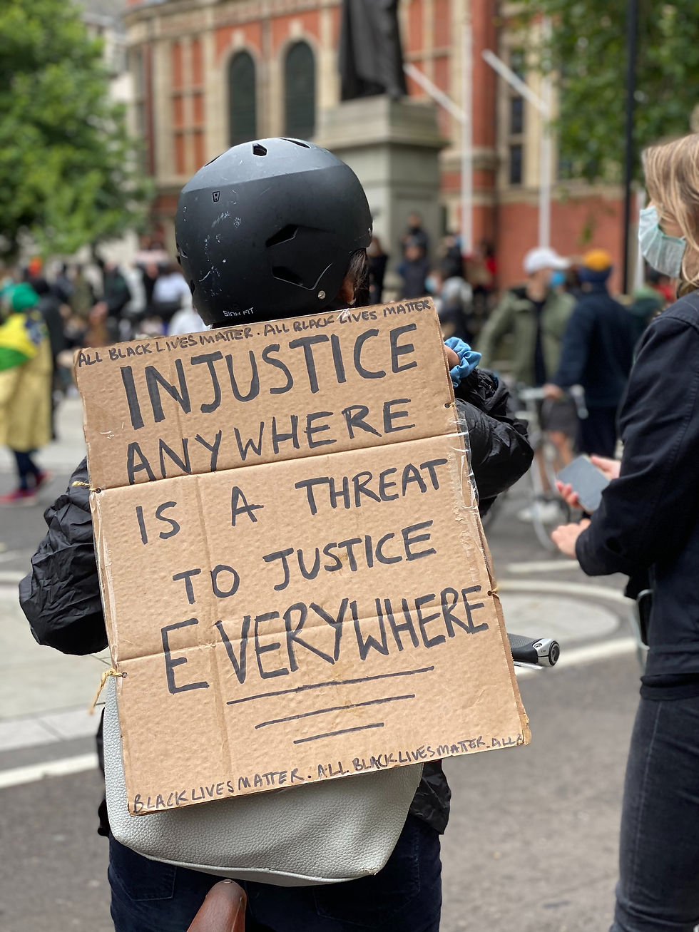 Photo of person carrying a cardboard sign on their back that reads, "Injustice anywhere is a threat to justice everywhere. All black lives matter."