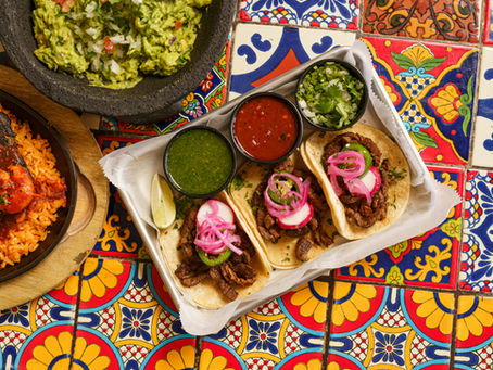 Tacos with pickled onions and sauces on vibrant patterned tiles, alongside guacamole and rice. Bright, festive setting.