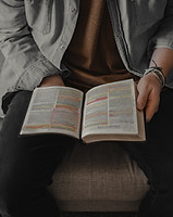 Man sitting on gray couch, holding an open bible in his lap.