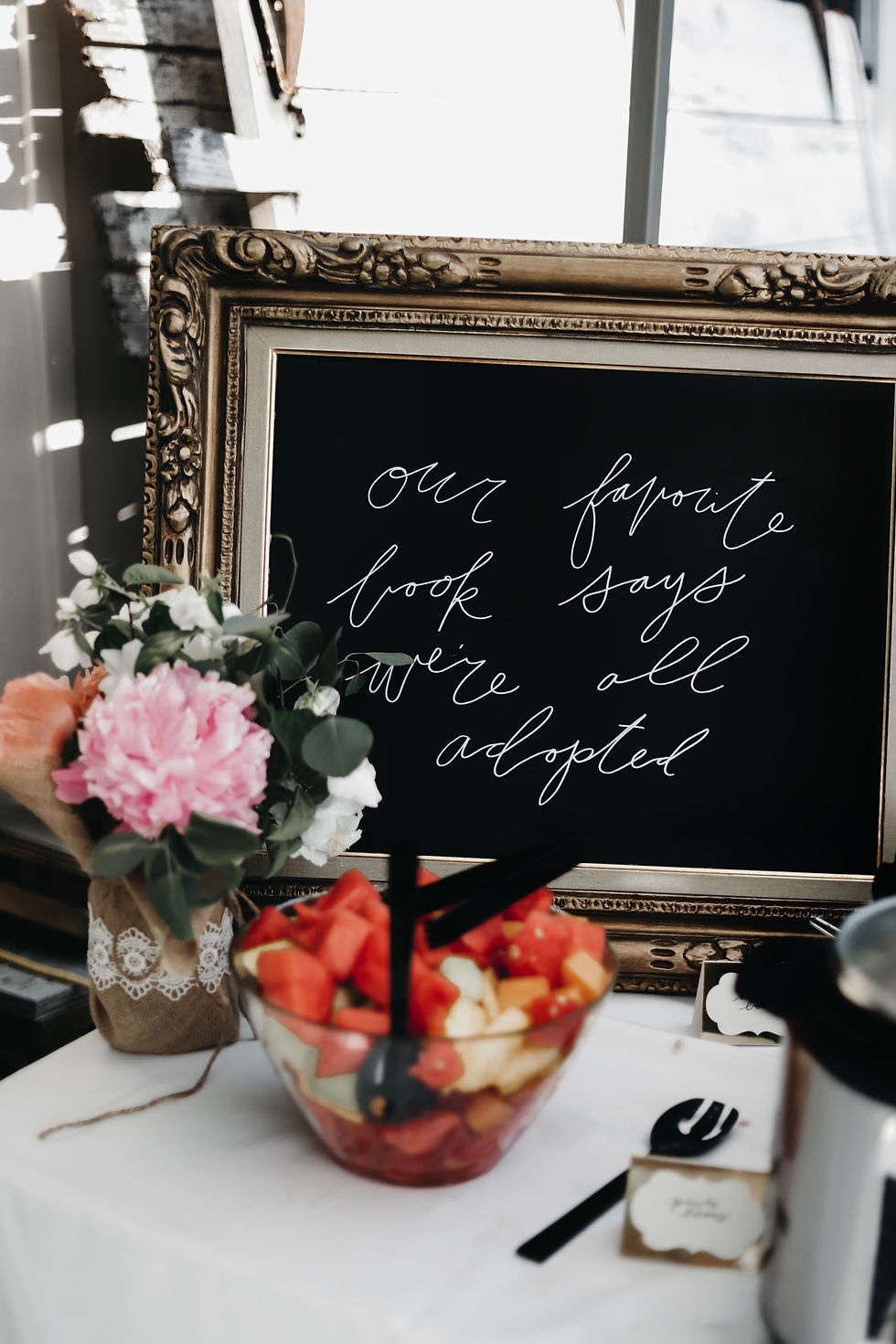 Elegant framed chalkboard reads "our favorite book says we're all adopted," beside flowers and fruit bowl on a white tablecloth.