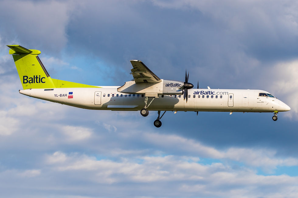 AirBaltic plane in flight, green tail, blue logo, against cloudy sky. Landing gear visible, propellers spinning, bright daylight.