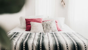 Bright bedroom with neatly made bed, striped blanket, pink and patterned pillows, and soft white curtains in a calm mood