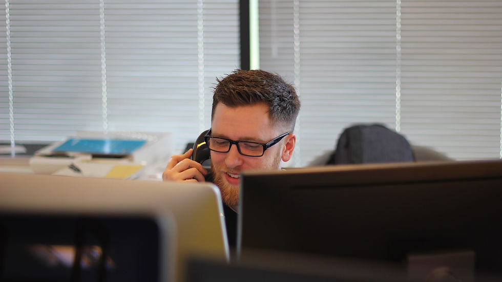Man wearing glasses talks on a phone at a desk between computer monitors in an office, smiling.