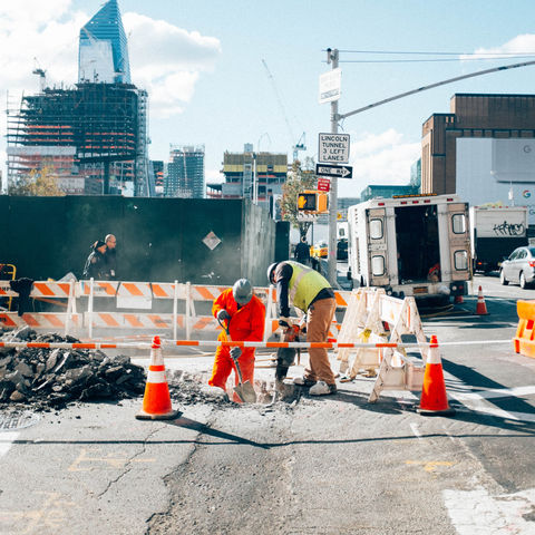 Construction workers repair a street trench at a busy NYC intersection, with Lincoln Tunnel signs, traffic, and skyline buildings.