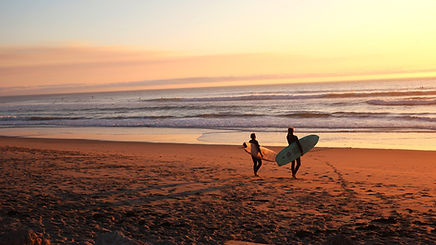 Two people walking with their surfboards on the beach towards the ocean during sunset