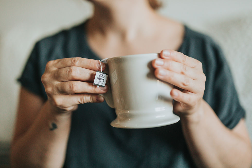 Hands holding a white mug with a tea bag labeled "Fonte". Person wearing a dark shirt. Calm setting with neutral background.