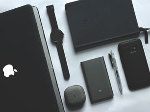 Masanın üzerinde bilgisayar, defter,saat, kalem, kulaklık, powerbank ve telefon