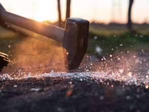 Close-up of a sledgehammer striking the ground, scattering debris with sunlight in the background, creating a dynamic, energetic scene.