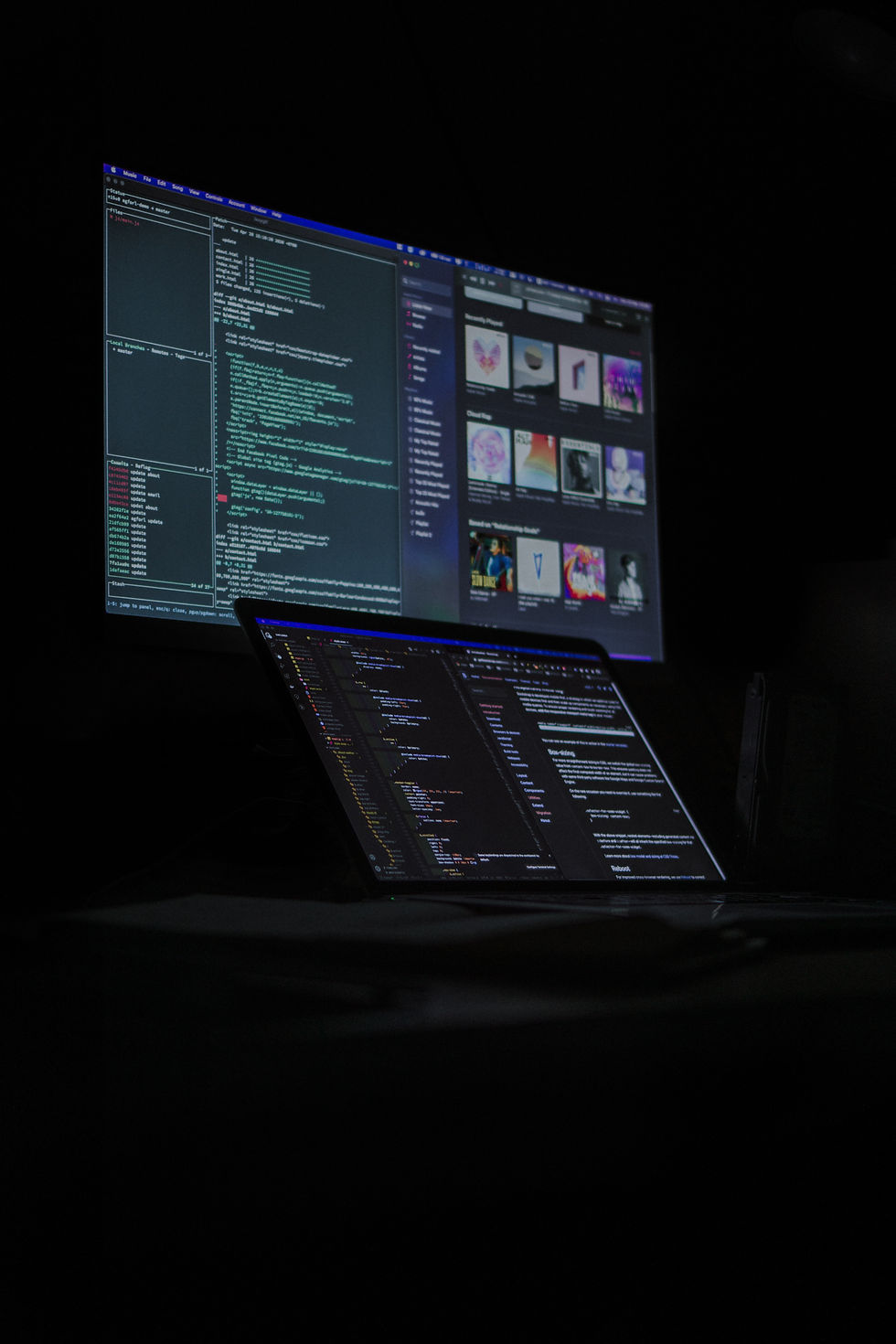 Dimly lit room with a monitor and laptop showing code and colorful images. Dark, tech-focused ambiance with visible text and programming setup.