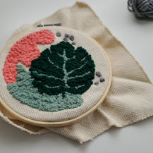Punch needle craft embroidery hoop with pink, green, and dark green abstract floral design on beige fabric, with a gray yarn ball in the background on a white surface.
