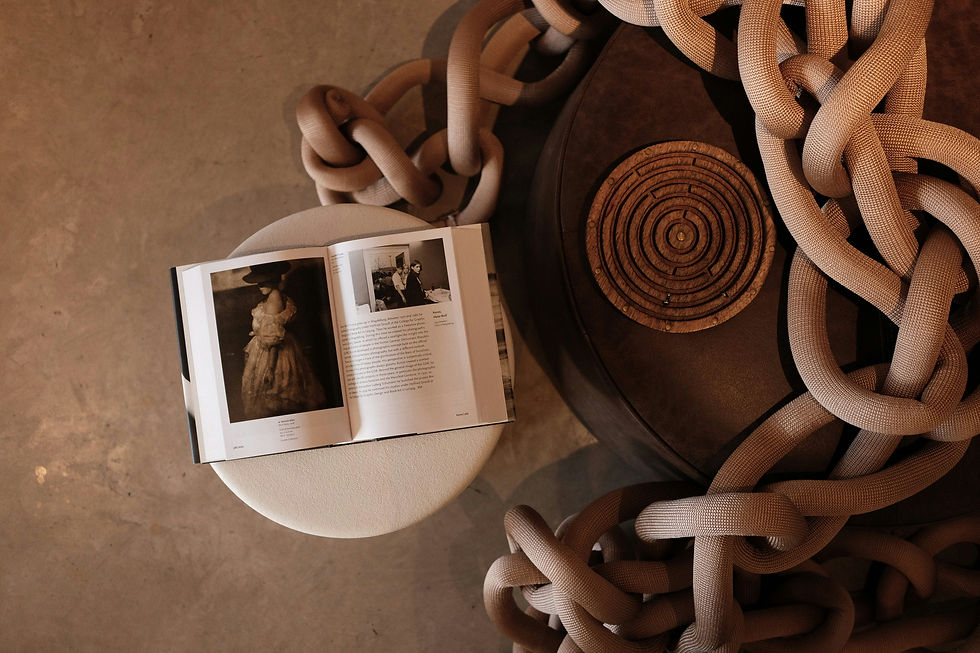 Open book on a round white stool, large knotted rope and embossed circular design on a brown cushion, set on a concrete floor.
