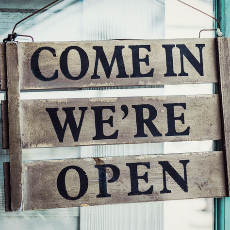 A rustic wooden sign hangs on a teal door, reading "COME IN WE'RE OPEN" in bold black letters, inviting and welcoming.