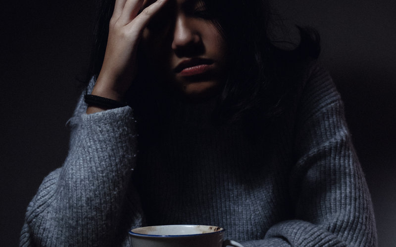 Young woman in a gray knit sweater holding a mug with her hand pressed to her forehead suggesting exhaustion or a headache