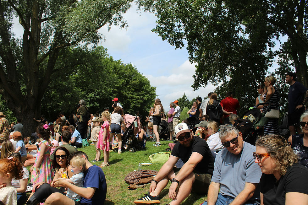 Menschen sitzen und stehen auf einer Wiese unter Bäumen bei einer sonnigen Parkveranstaltung.