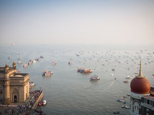Hazy Mumbai harbor with the Gateway of India, Taj Mahal Palace dome, and many boats and ferries on calm water