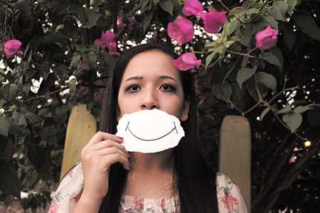 A person is holding up a piece of paper with a smile drawn on it, in front of their mouth