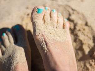 Sandy feet with blue toenails on a beach, immersed in soft, golden sand, evoking a relaxed and carefree beach vibe.