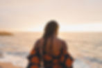 Woman in a vibrant dress with orange patterns stands by the ocean at sunset, gazing at the horizon. Calm and serene atmosphere.
