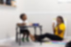 Boy sitting at the table and woman sitting on the ground teaching him. Both are smiling. 