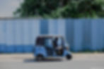 Man drives a blue tuk-tuk along a quiet street beside a tall corrugated metal fence and leafy trees.